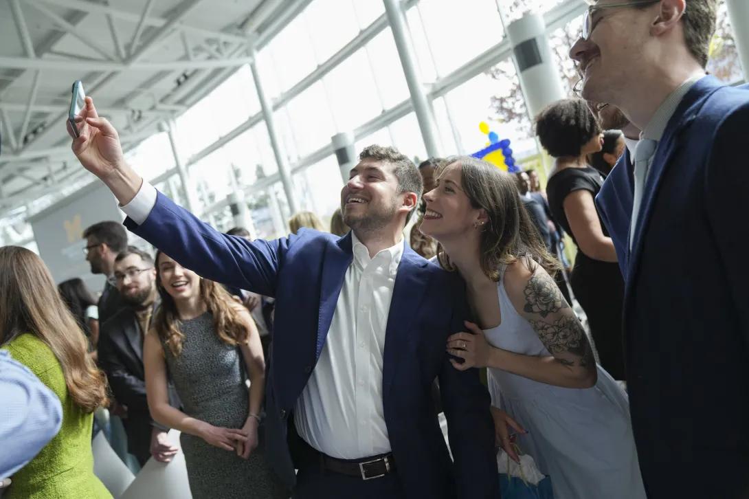 In a bright, crowded atrium during a celebration, a person in a blue suit holds up a phone to take a selfie with a companion in a light dress, while other guests mingle nearby.