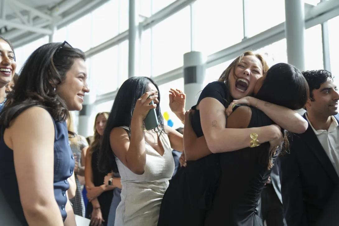  In a sunlit atrium during a celebration, two attendees hug tightly while friends nearby smile and record the moment on a phone.