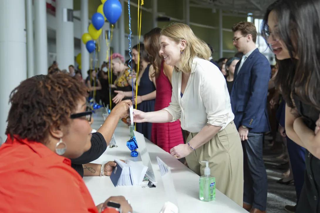At a busy Match Day check-in table decorated with maize-and-blue balloons, a staff member hands an envelope to an attendee while others wait in line; hand sanitizer and stacks of envelopes sit on the table.