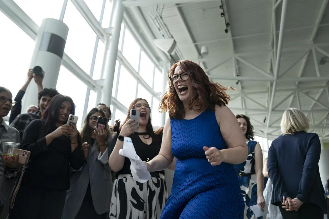 In a bright atrium filled with cheering onlookers holding up phones, a person in a blue dress steps forward holding a sheet of paper, as if reacting to exciting news during a celebration.