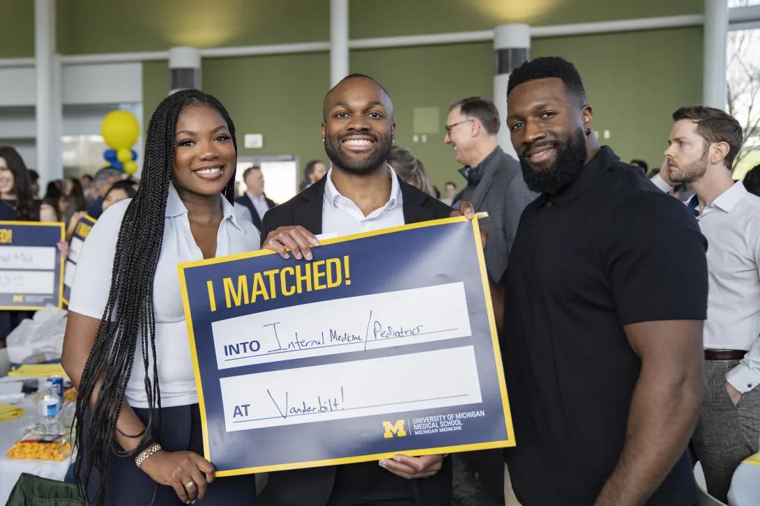 Three attendees pose at a crowded University of Michigan Medical School Match Day event holding an “I MATCHED!” sign that reads “Internal Medicine/Pediatrics at Vanderbilt,” with maize-and-blue balloons and other guests in the background.