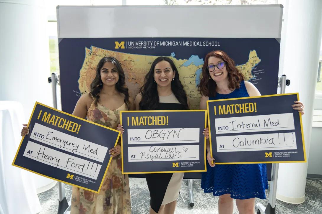 Three medical students pose in front of a University of Michigan Medical School U.S. map display holding “I MATCHED!” signs announcing matches in Emergency Medicine at Henry Ford, OB/GYN at Corewell–Royal Oak, and Internal Medicine at Columbia.