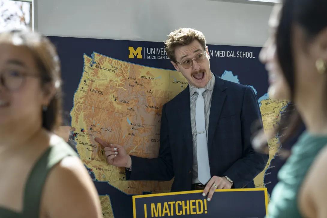 At a University of Michigan Medical School Match Day display map of the United States, a person in a suit points to California while holding an “I MATCHED!” sign.