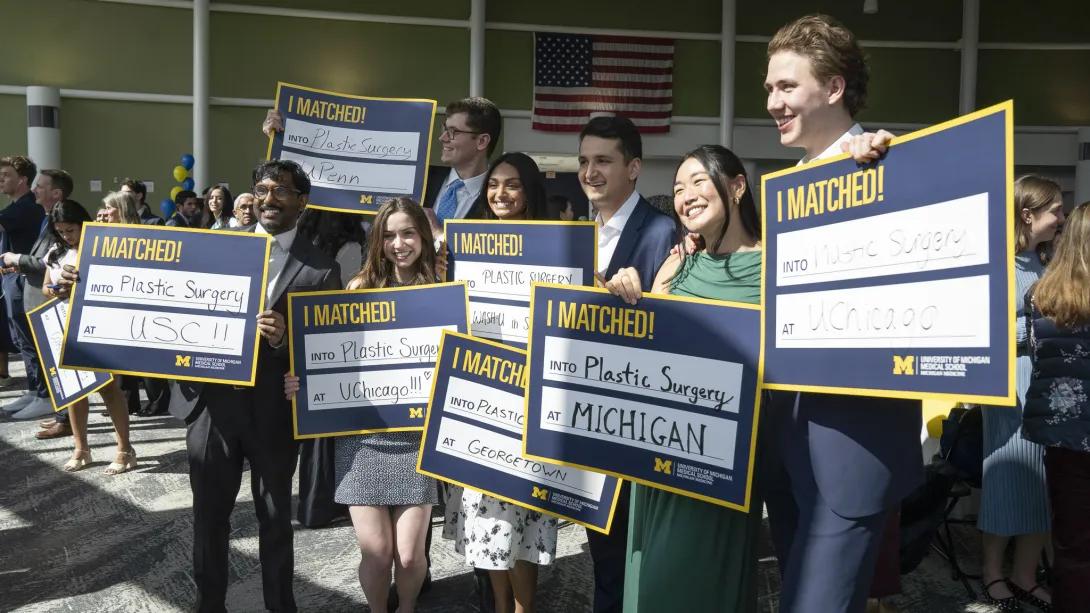 A group of medical students pose at a Match Day celebration holding “I MATCHED!” signs for plastic surgery programs at USC, Penn, WashU in St. Louis, Georgetown, Michigan, and the University of Chicago, with other attendees and a U.S. flag in the background.