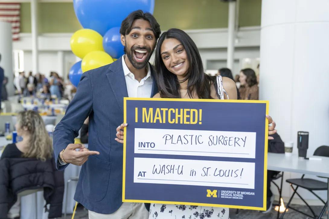 Two people stand together at a celebratory event holding a University of Michigan Medical School sign that reads, “I MATCHED! Into Plastic Surgery at Wash-U in St. Louis!” with blue and yellow balloons and seated guests in the background.