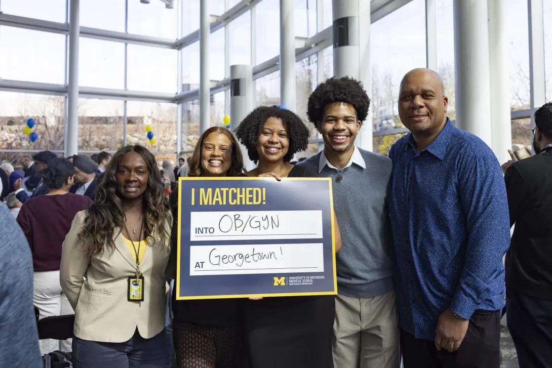 A group of five people pose at a crowded Match Day celebration in a bright glass lobby, holding a University of Michigan Medical School sign that reads, “I MATCHED! into OB/GYN at Georgetown!”