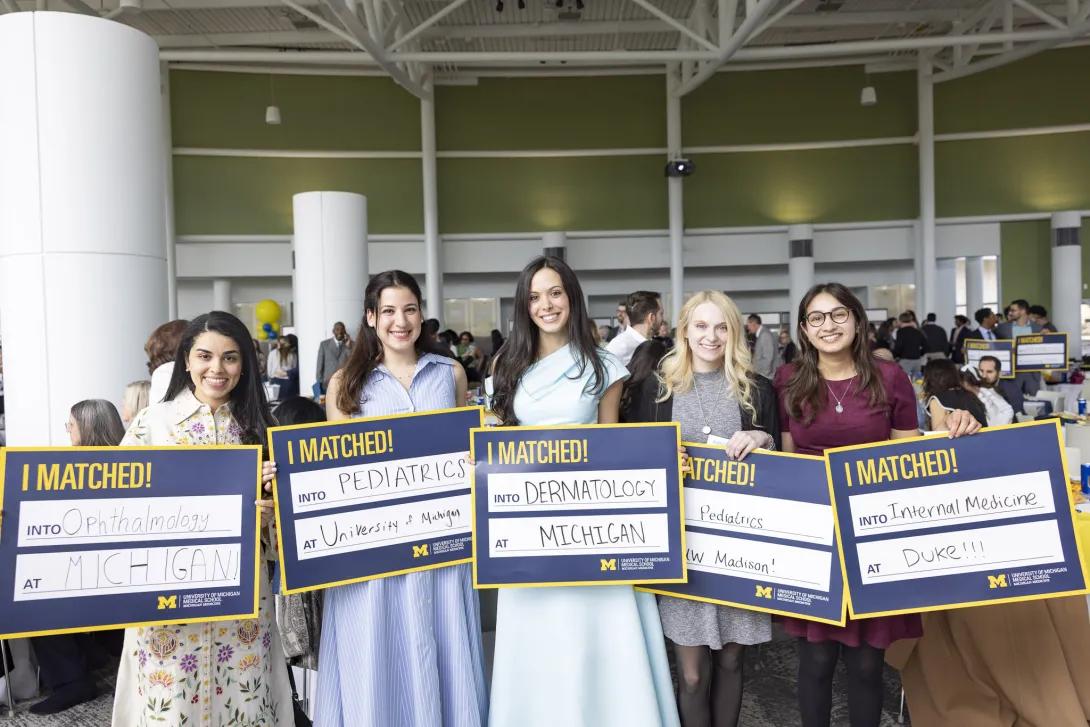 Five medical students stand together at a crowded Match Day celebration holding “I MATCHED!” signs for Ophthalmology at Michigan, Pediatrics at the University of Michigan, Dermatology at Michigan, Pediatrics at UW–Madison, and Internal Medicine at Duke.