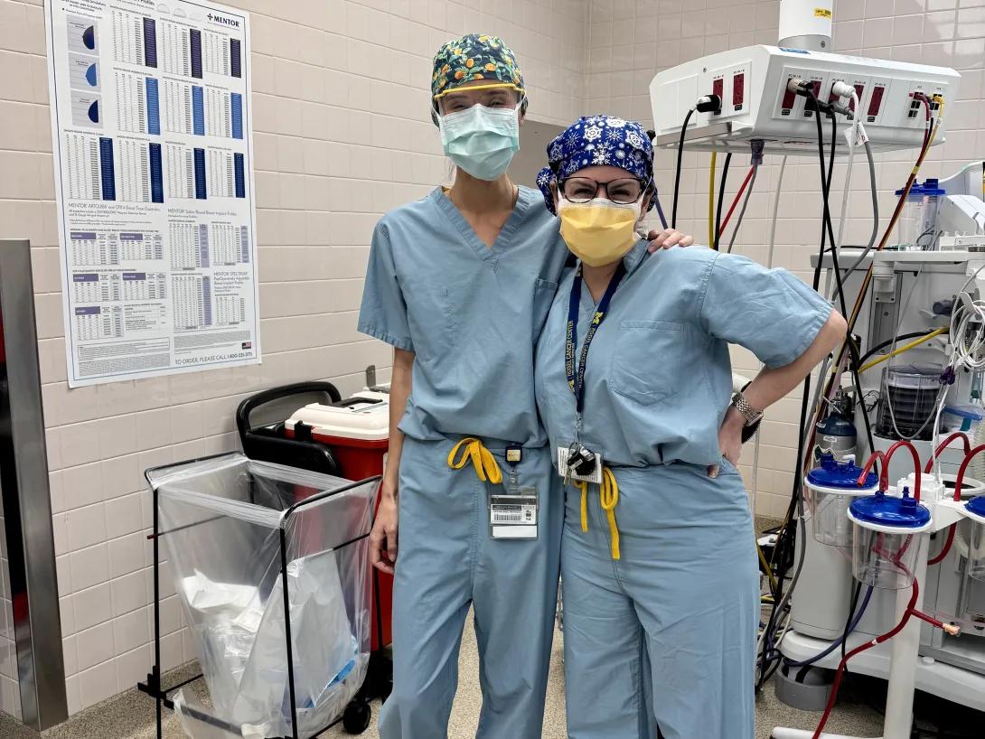 Two people in light-blue surgical scrubs and patterned caps stand side by side in a hospital operating room, one with an arm around the other. Medical equipment, tubing, and wall-mounted outlets are visible behind them, along with a poster and a trash/linen bin.