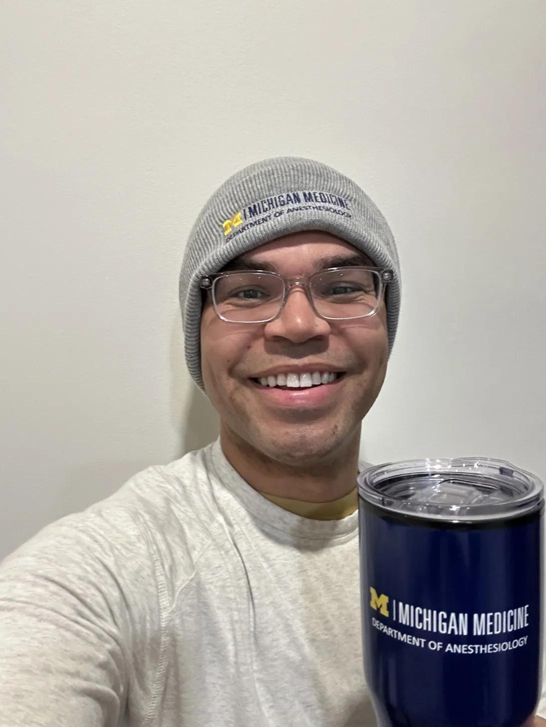 A person takes a selfie against a plain wall, wearing a gray knit beanie with “Michigan Medicine” text and holding a navy insulated tumbler labeled “Michigan Medicine Department of Anesthesiology."”