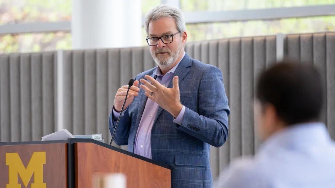 A speaker gestures while presenting at a University of Michigan podium during the Department of Anesthesiology Research Retreat.