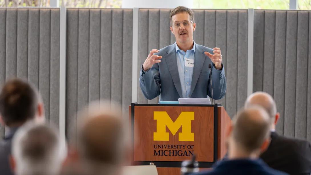 A speaker gestures while presenting from a University of Michigan podium to attendees at the Department of Anesthesiology Research Retreat.