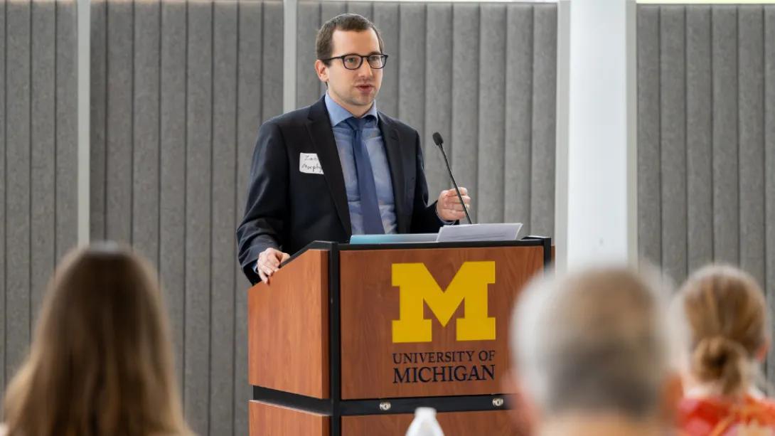 A presenter speaks at a University of Michigan podium while audience members listen during the Department of Anesthesiology Research Retreat.