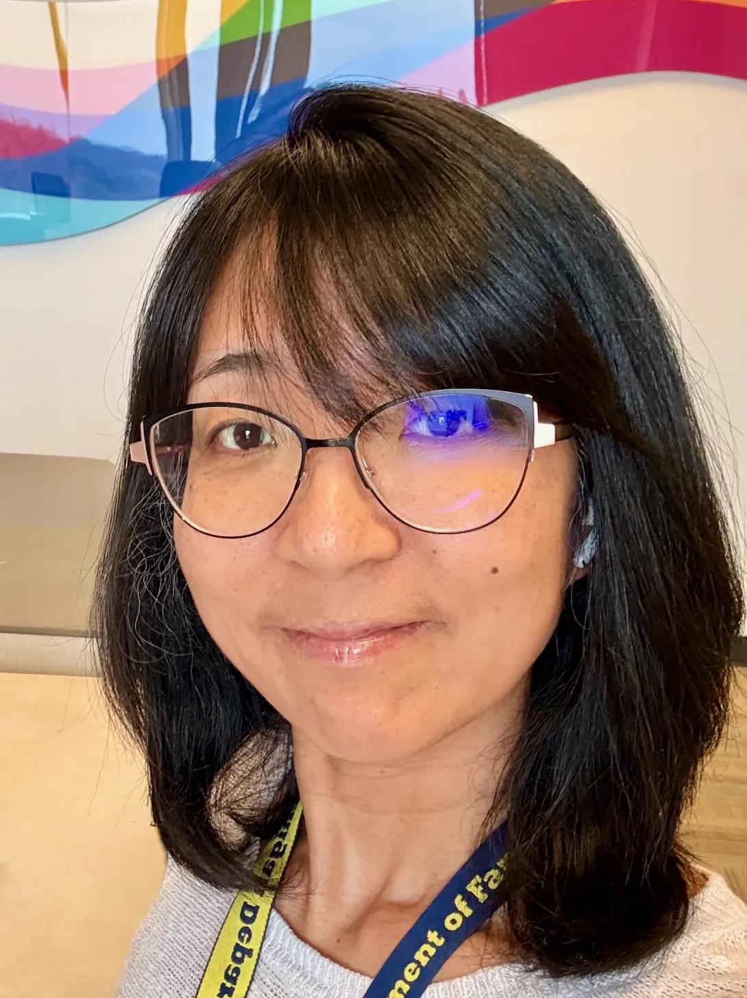 Asian woman with shoulder length dark hair and glasses, wearing a light grey sweater.