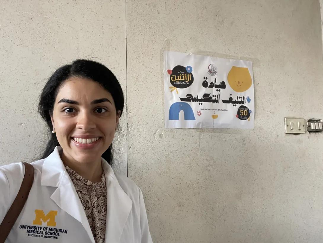 A person wearing a University of Michigan Medical School white coat smiles for a selfie beside a health education poster with Arabic text on a wall.