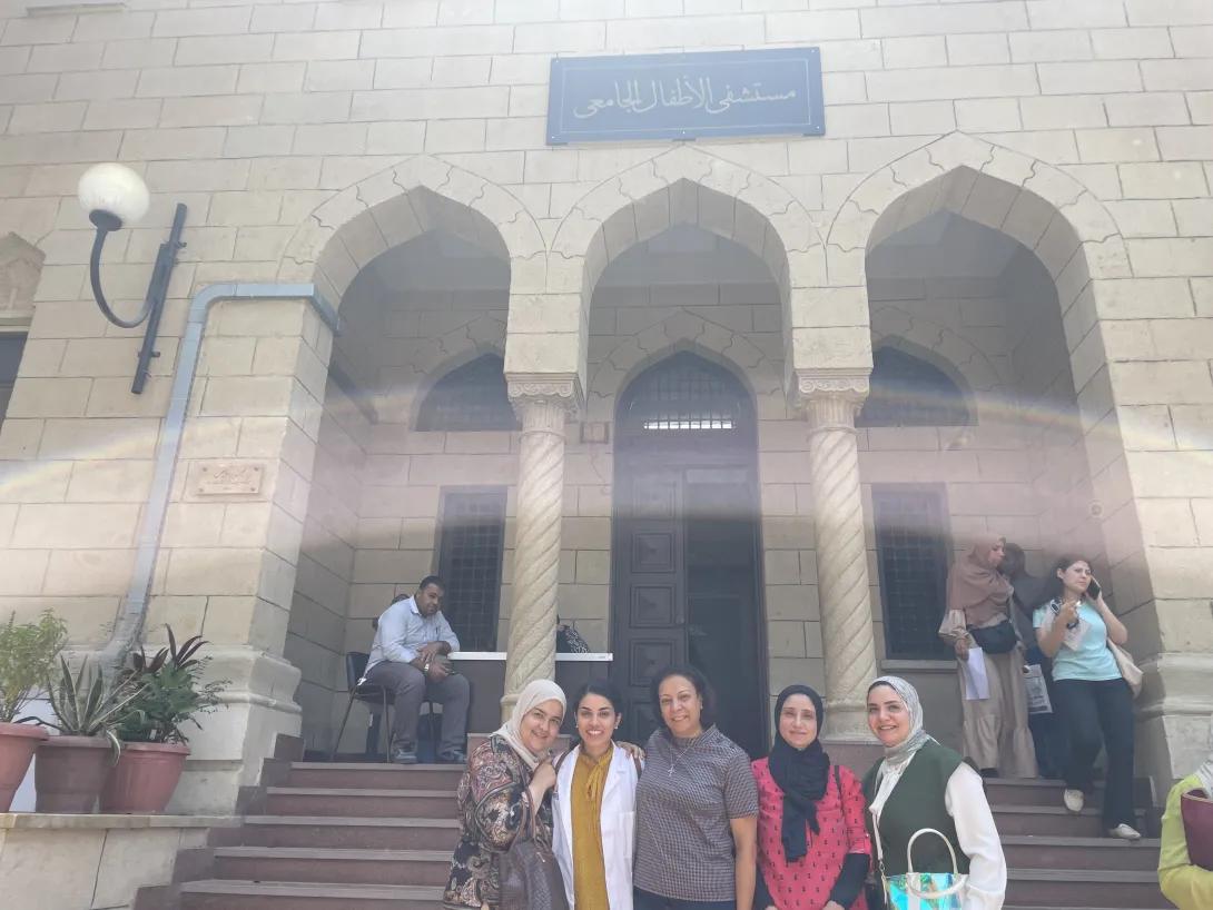 A group of five people pose on the steps outside a stone building with arched doorways and an Arabic sign above the entrance. One person wears a University of Michigan white coat, and sunlight creates a bright lens flare across the photo.