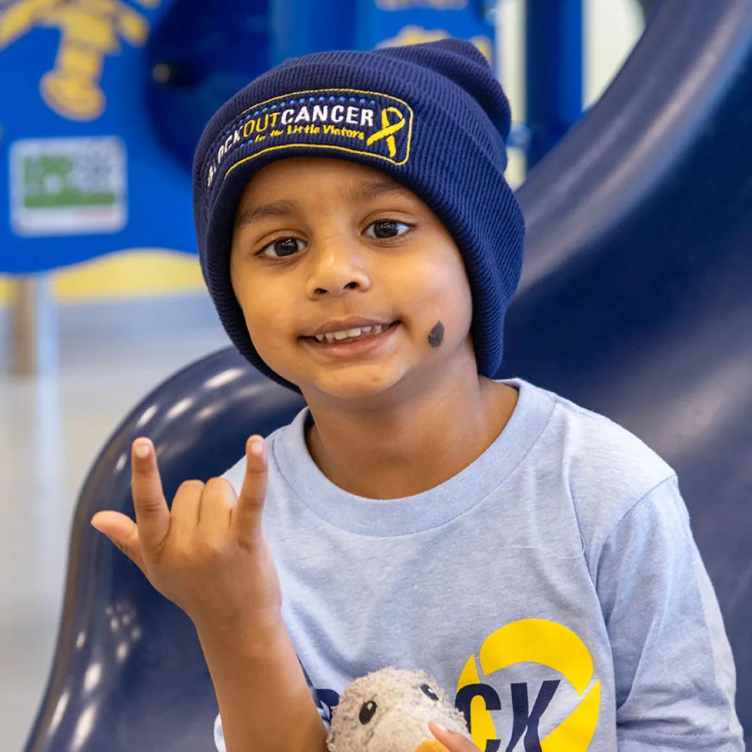 A smiling child in a Block Out Cancer t-shirt and hat sits at the bottom of a slide and makes a playful hand gesture.