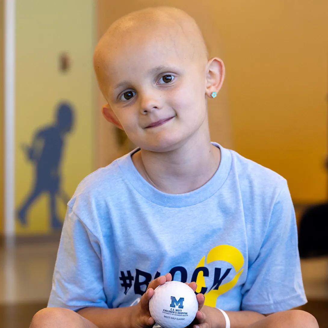 A child wearing a Block Out Cancer t-shirt smiles and holds a small white ball with a block M.