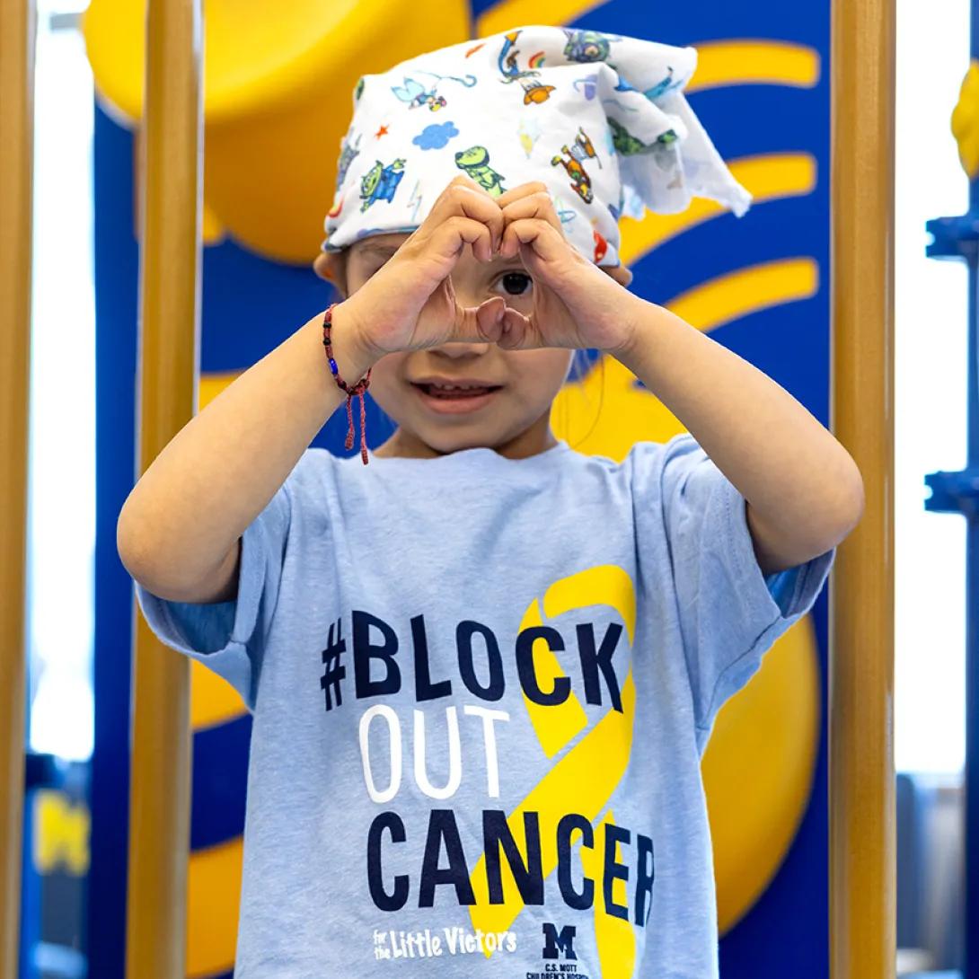 A child wearing a Block Out Cancer t-shirt holds up her hands in a heart gesture.