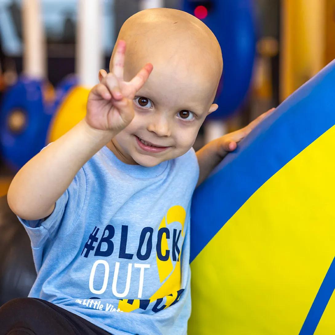 A smiling child wearing a Block Out Cancer t-shirt holds up their hand in a peace sign.
