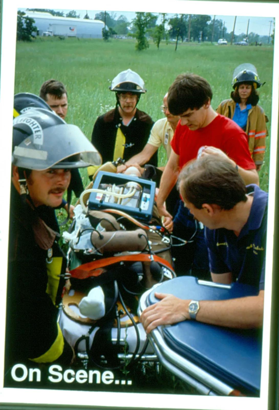 Group0 of first responders all surrounding a stretcher outside in a field. Text on the image says "on scene"